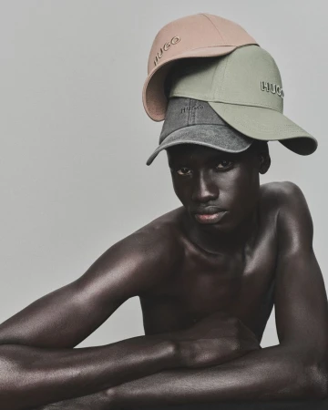 Dark-skinned adult male with three layered caps from HUGO fashion label, posing in a close-up studio shot with neutral background.