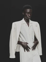 Black man wearing a structured white suit jacket over matching shirt against a dark background.