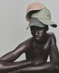 Shirtless male model wearing three stacked baseball caps with HUGO branding against a plain neutral background.