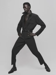 Black man wearing a dark jumpsuit and dress shoes posing against a plain grey studio backdrop for HERO Magazine HUGO project.