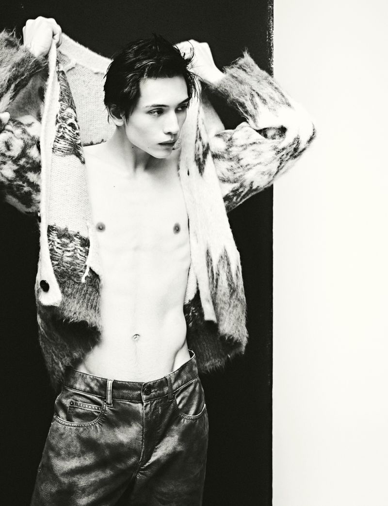 Monochrome studio fashion shot of a shirtless slender adult male adjusting a patterned sweater, wearing shiny pants against a dual backdrop.