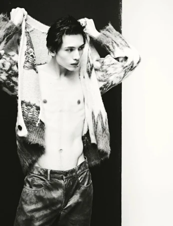 Monochrome studio fashion shot of a shirtless slender adult male adjusting a patterned sweater, wearing shiny pants against a dual backdrop.