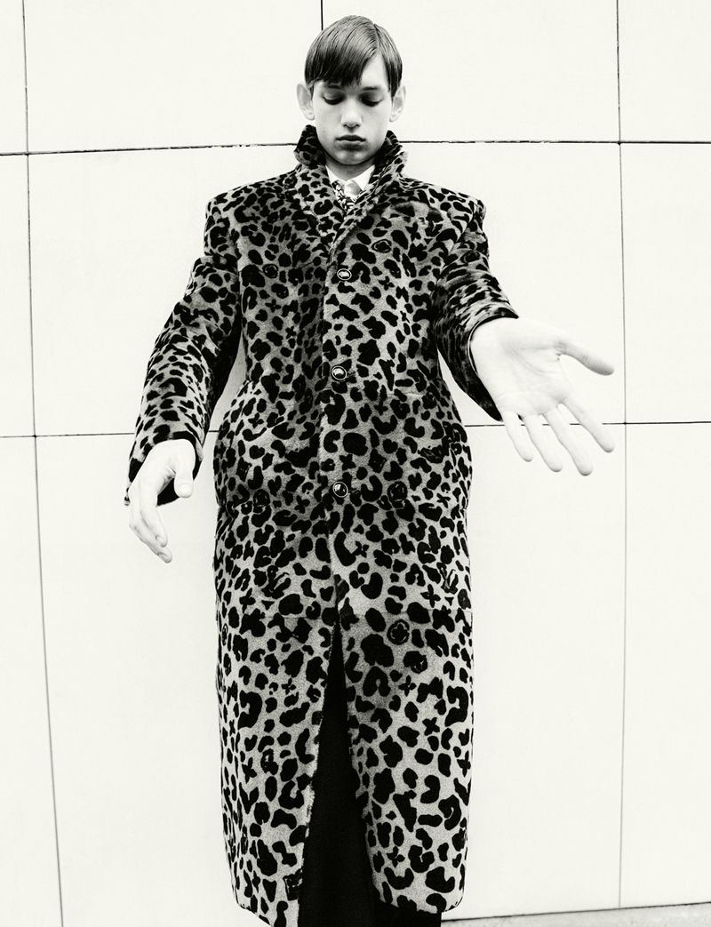 A fair-skinned young adult is photographed indoors wearing a leopard print coat, staring down with arms slightly extended in monochrome.