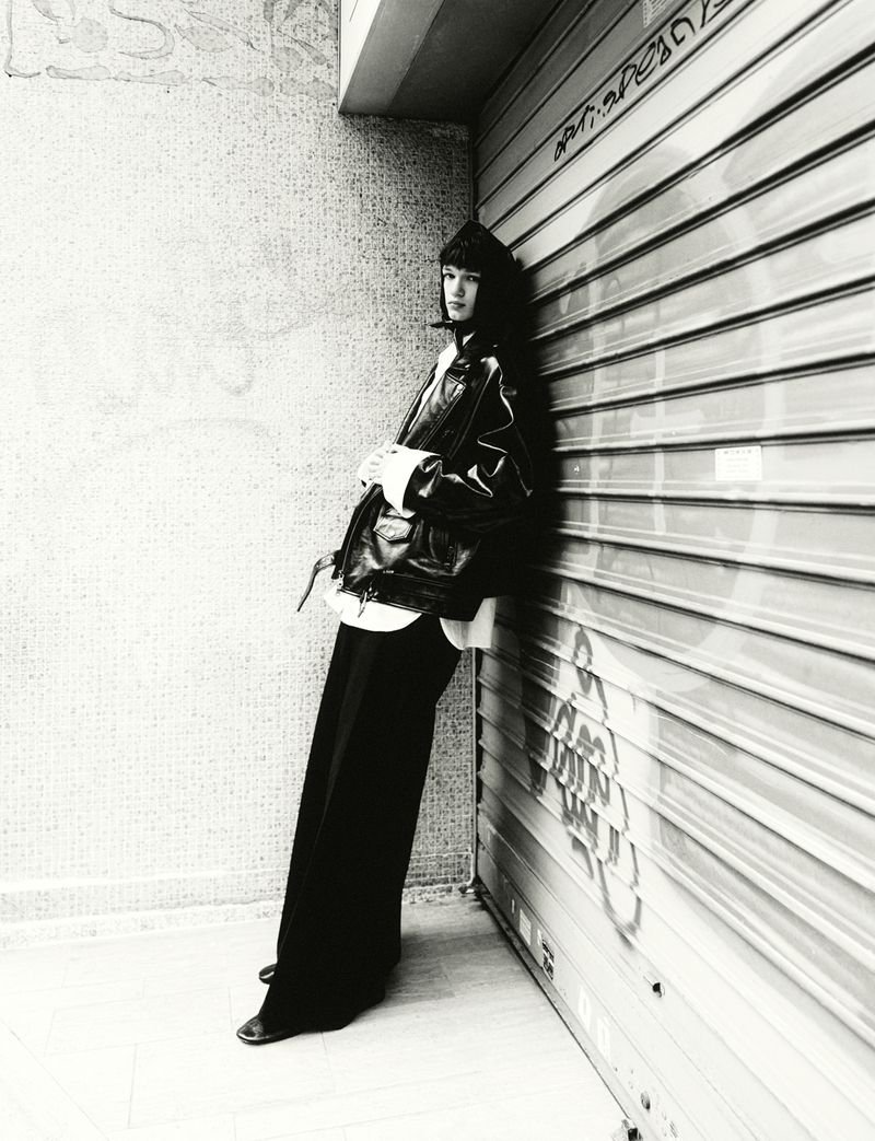 Woman in black jacket and trousers leans against a shuttered metal wall in a black and white fashion photograph taken outdoors.