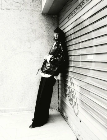 Woman in black jacket and trousers leans against a shuttered metal wall in a black and white fashion photograph taken outdoors.