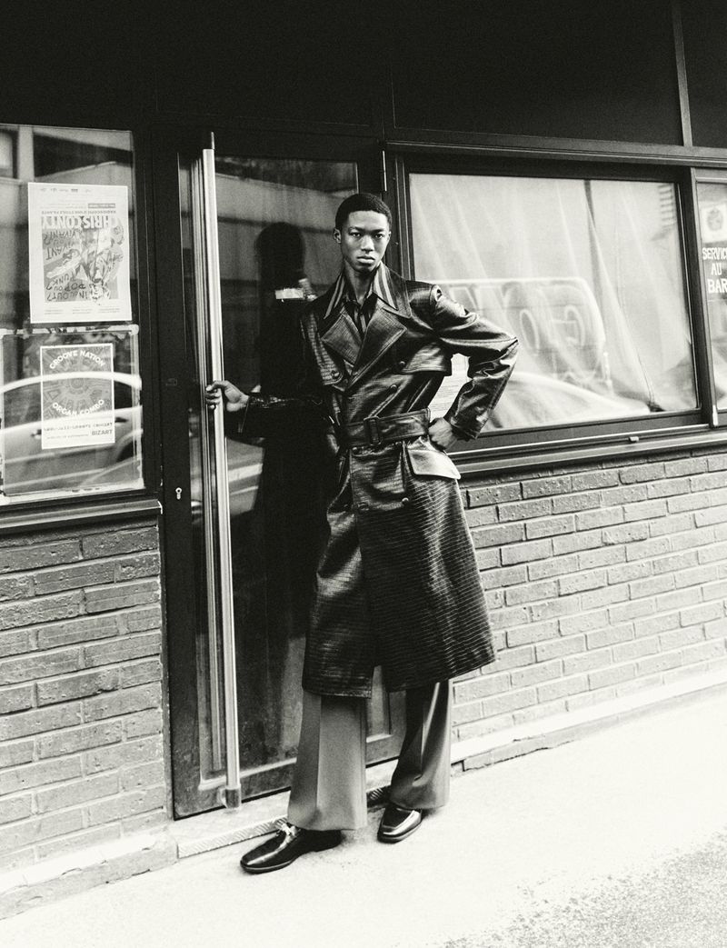 A solitary African-descent adult model stands confidently outside a shop entrance, wearing a distinctive black leather trench coat fashion.