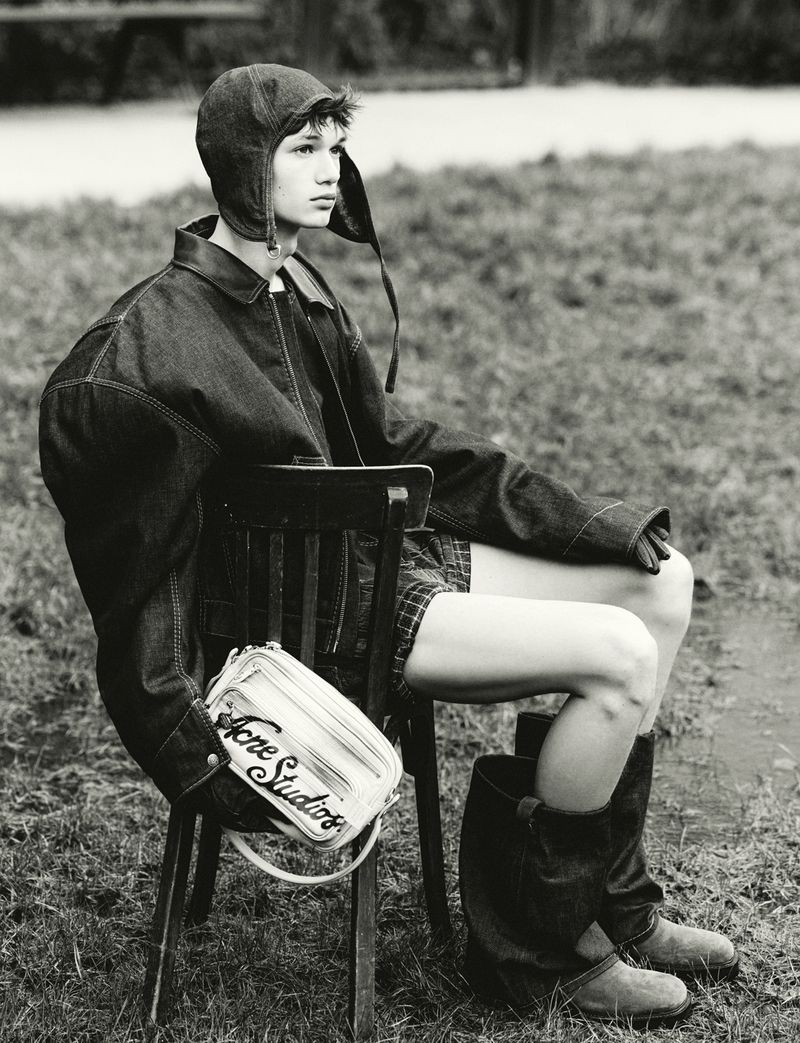 Monochrome fashion portrait of a model wearing an aviator hat, oversized denim jacket, and boots, seated outdoors on a chair.