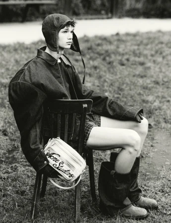 Monochrome fashion portrait of a model wearing an aviator hat, oversized denim jacket, and boots, seated outdoors on a chair.