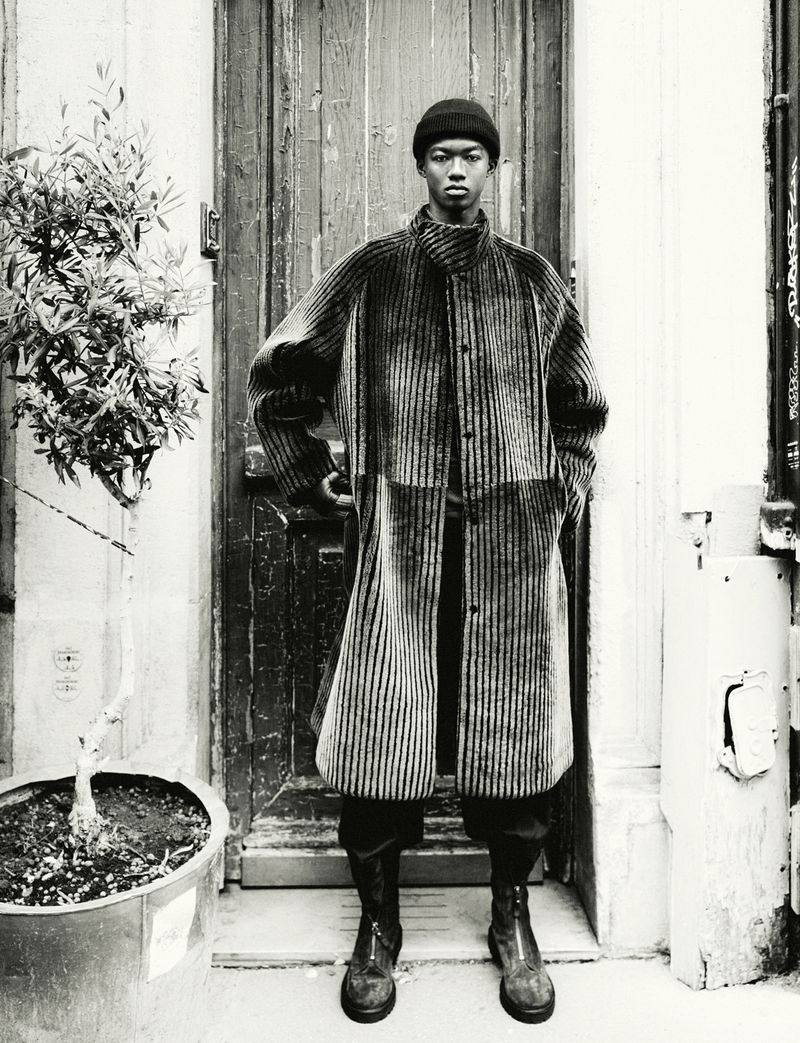 Fashion shot of a black male in a striped coat, hat, and boots, standing in front of a weathered wooden door with potted plant.