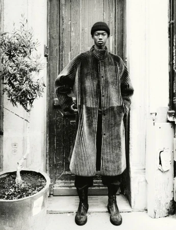 Fashion shot of a black male in a striped coat, hat, and boots, standing in front of a weathered wooden door with potted plant.