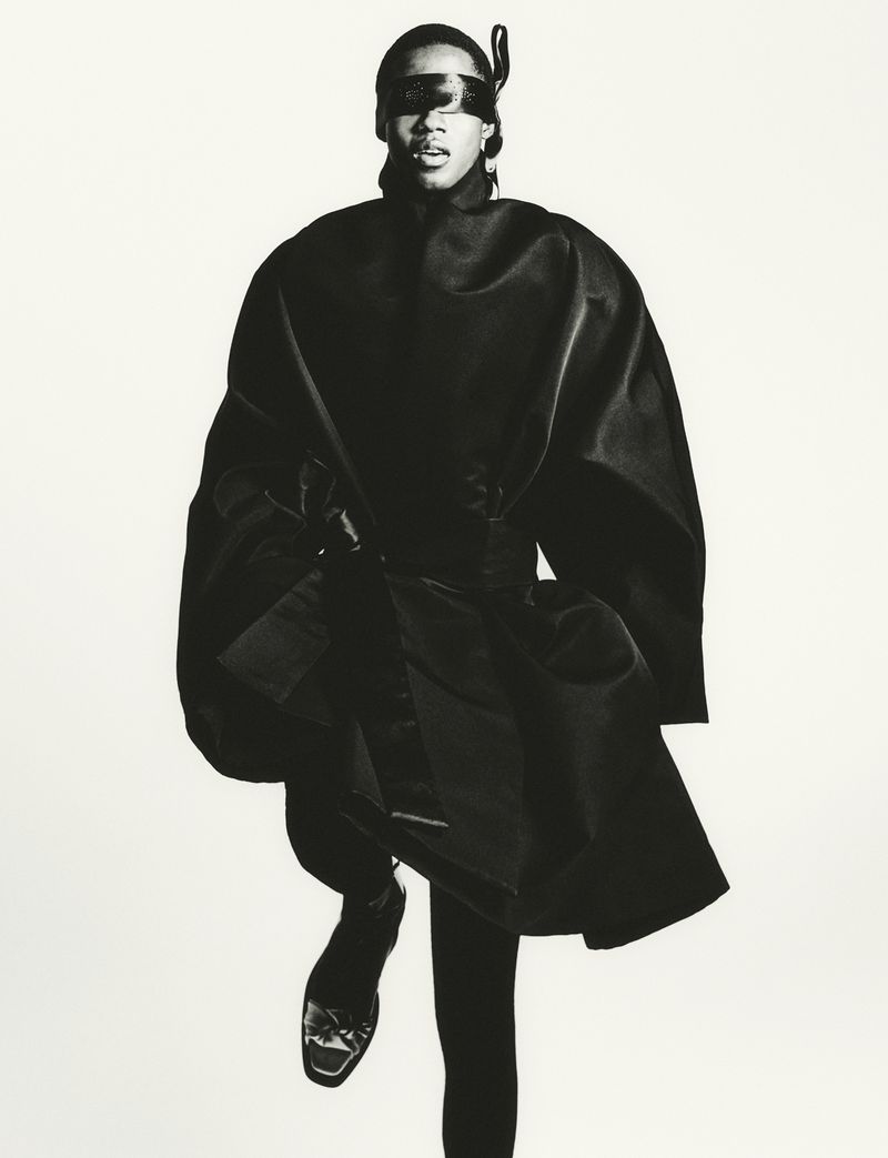 A single adult female model in a dark avant-garde fashion look poses in a full shot against a bright white studio backdrop.