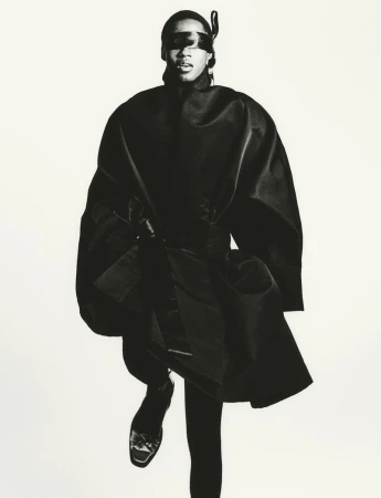 A single adult female model in a dark avant-garde fashion look poses in a full shot against a bright white studio backdrop.