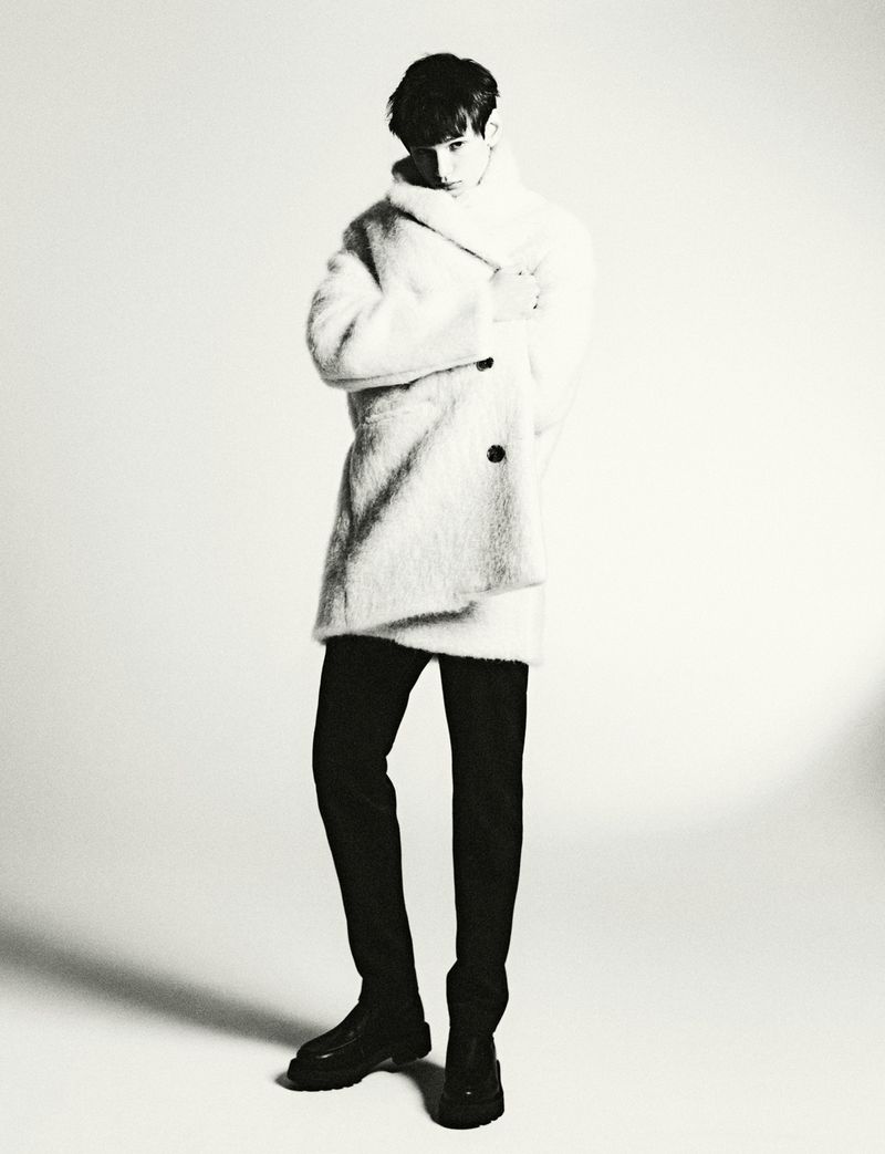 Model wearing a textured white coat and dark trousers posing against a bright plain background for HERO Magazine.