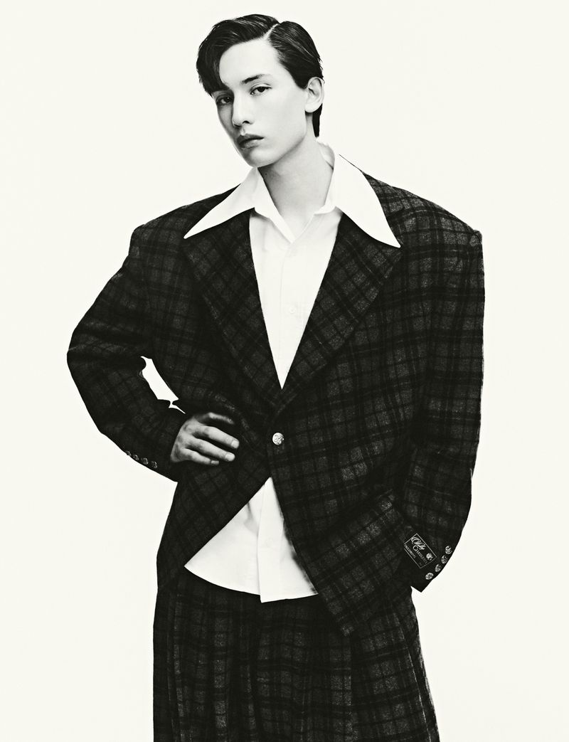 Monochrome studio shot of a slender young adult with short dark hair wearing a plaid suit with a white collared shirt.
