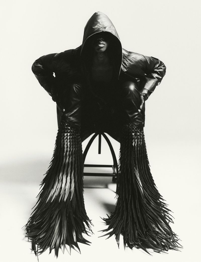 Model in a black hooded leather jacket seated on a stool against a white seamless background