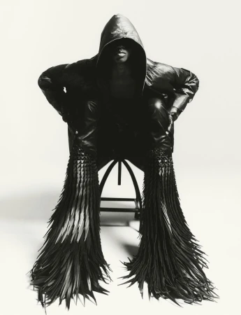 Model in a black hooded leather jacket seated on a stool against a white seamless background