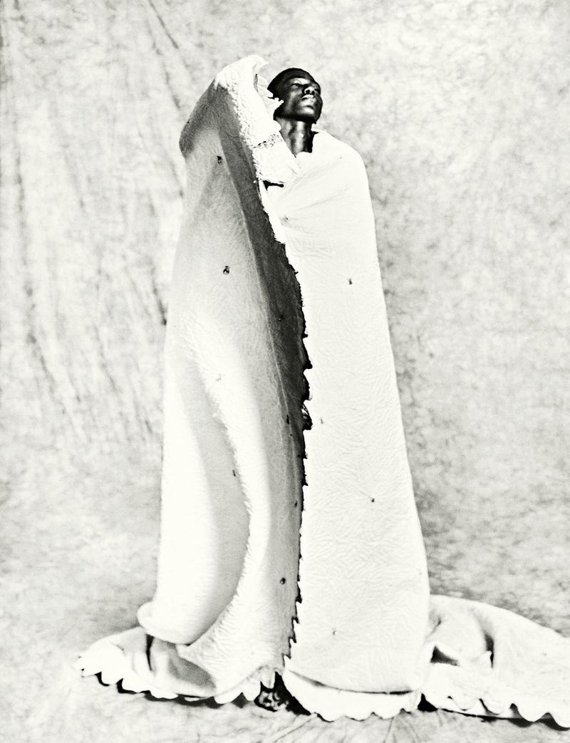 Portrait of black man wrapped in textured white fabric against muslin backdrop in studio, captured in black and white. 