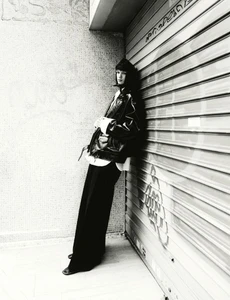 Monochrome fashion editorial image featuring a model leaning against a shuttered storefront and textured concrete wall.