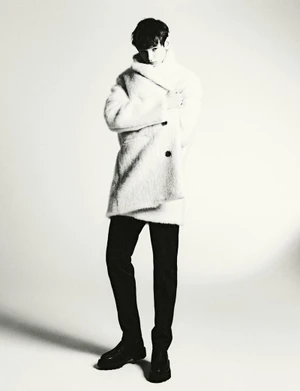 Model wearing a textured white coat and dark trousers posing against a bright plain background for HERO Magazine.