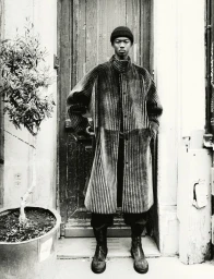 Black man wearing a textured long coat and beanie posing in front of a wooden doorway with a potted plant.