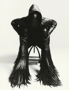 Model in a black hooded leather jacket seated on a stool against a white seamless background