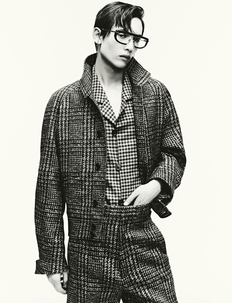 Young male model wearing thick glasses and a plaid jacket against a plain background