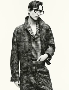 Young male model wearing thick glasses and a plaid jacket against a plain background