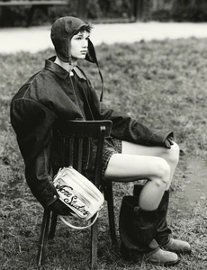 Monochrome fashion portrait of a model wearing an aviator hat, oversized denim jacket, and boots, seated outdoors on a chair.