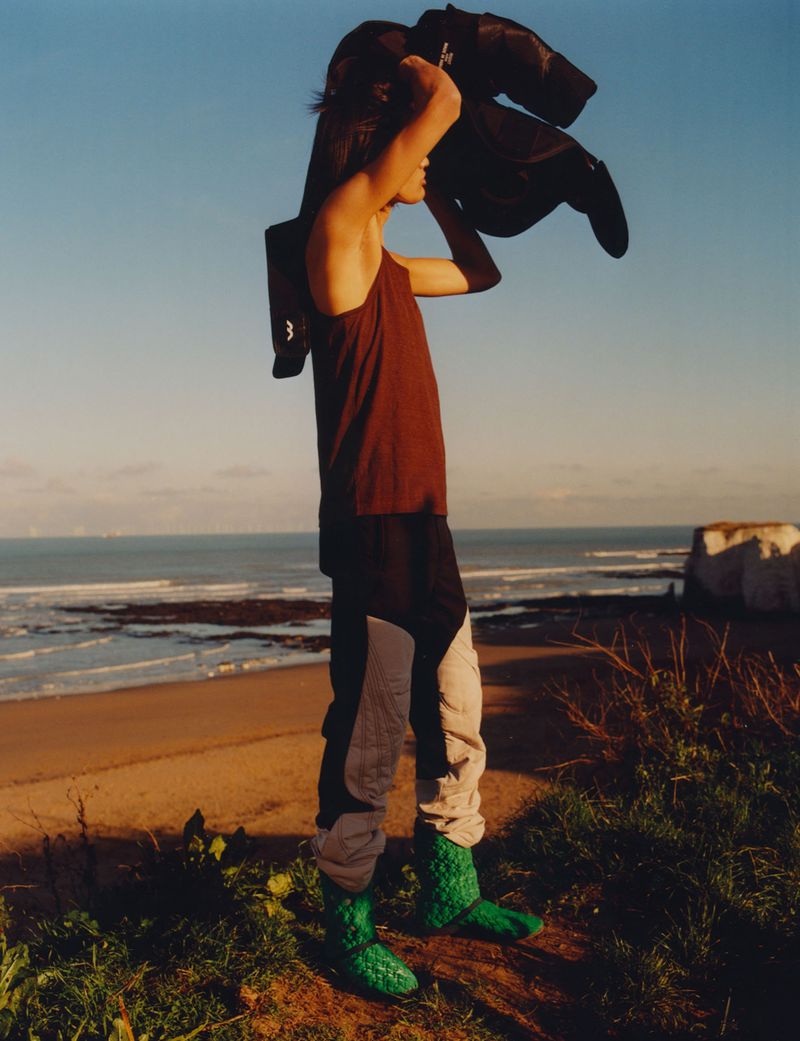 Person poses in rust tank top, color-block pants and green boots on sandy coastline with jacket over shoulder on sunny day.