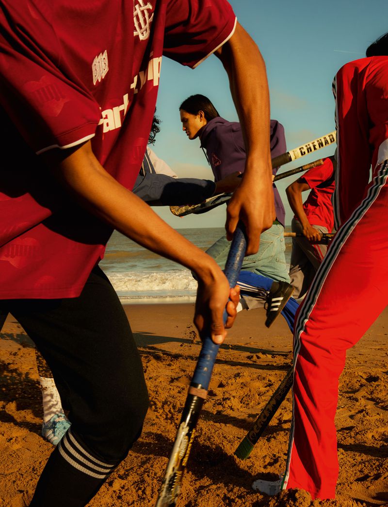 Group of people playing hockey on a sandy beach near ocean with blue sky, wearing athletic clothing, red and blue tones, medium shot.