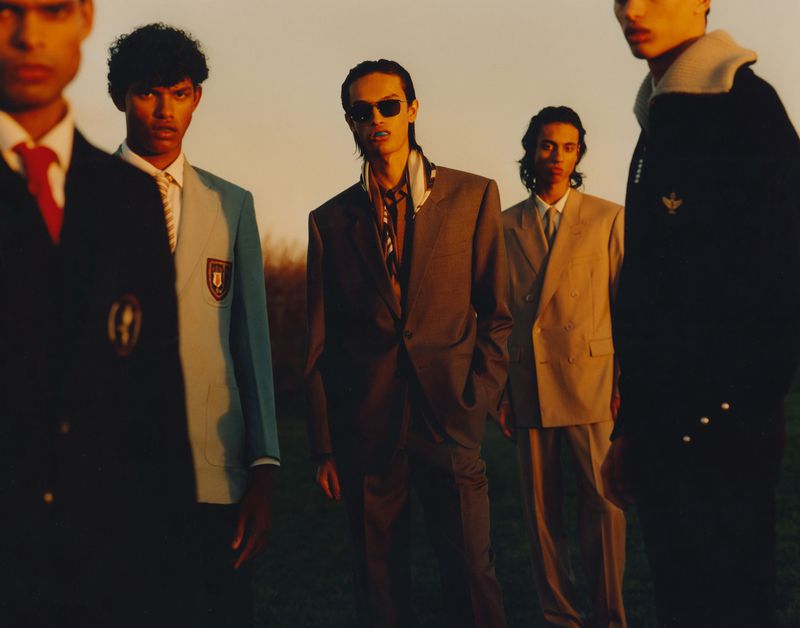 Five male fashion models in suits and jackets posing outdoors against a golden-hour sky in a group fashion shot with a shallow depth of field.