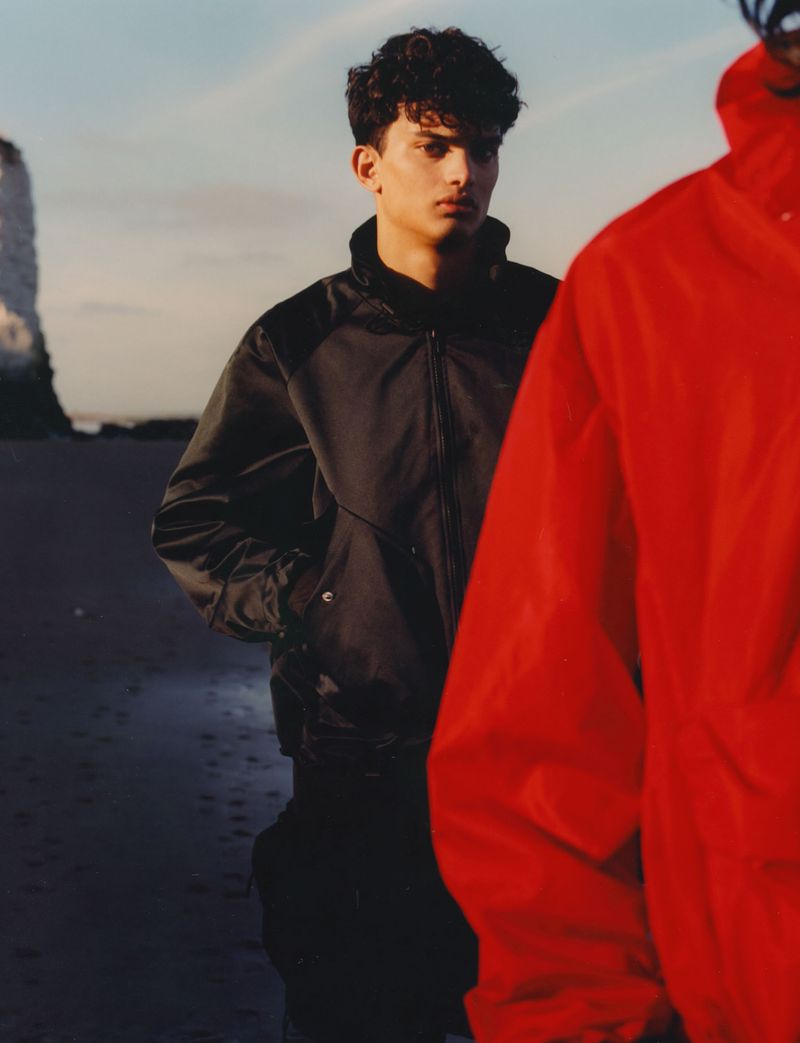 Two people stand on a sandy beach with a rocky cliffside in the background, one in a red jacket and one in a dark jacket.