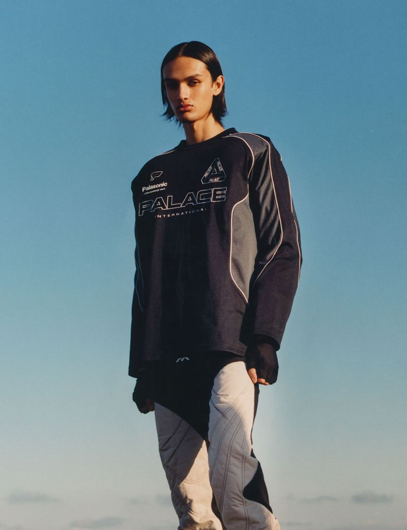 Fashion portrait of a young adult male in a Palace International long-sleeve shirt against a light blue sky backdrop.