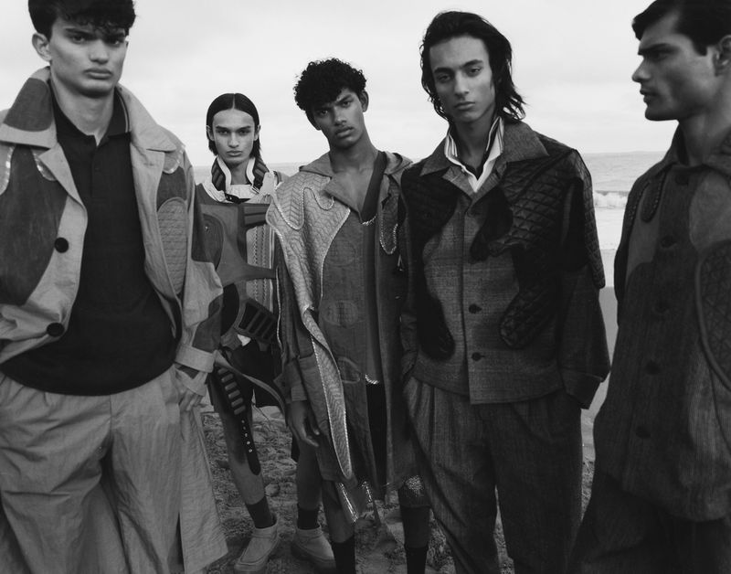 Monochrome fashion shot of five slender young adult models on a beach wearing avant-garde patterned jackets and neutral colored pants and footwear.
