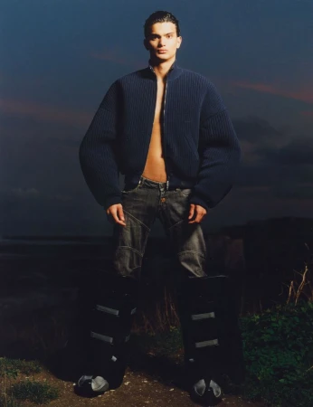 Male fashion model poses outdoors wearing a zip-up bomber jacket, distressed jeans, and futuristic boots against a twilight sky.