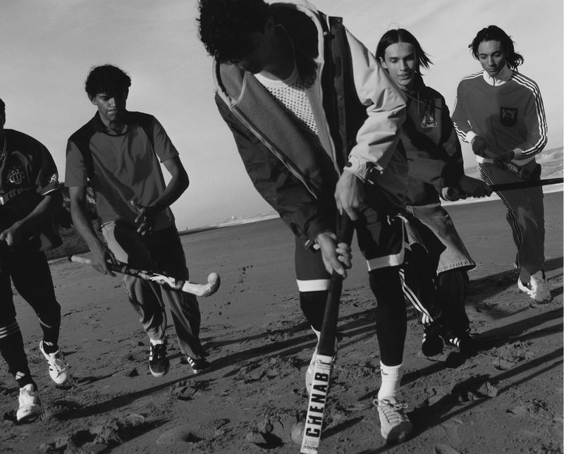 Black and white photo of a group of young adult male field hockey players in sportswear holding their hockey sticks on a beach.