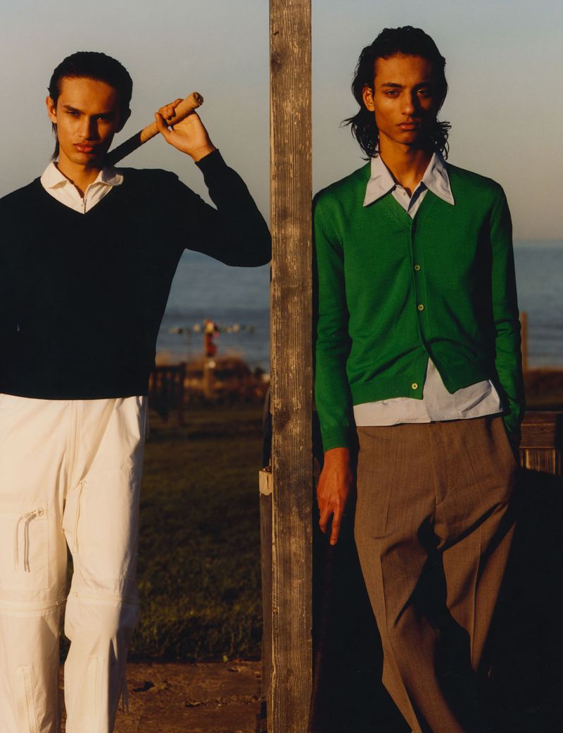 Two adult men in vintage-inspired fashion stand outdoors near a wooden post, one holding a polo mallet in a bright, sunlit scene.