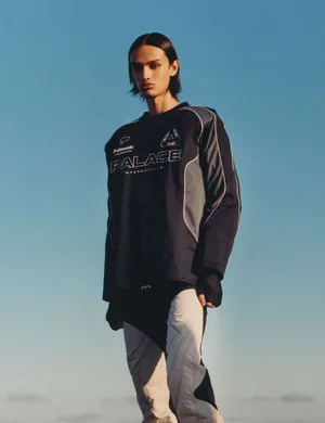 Model wearing a dark long-sleeve athletic jersey and white trousers against a clear blue sky background