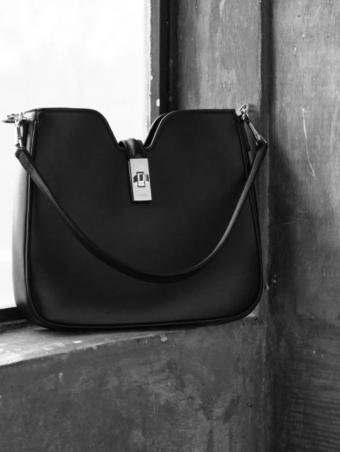 Elegant black leather shoulder bag with a silver clasp, displayed on a textured concrete surface near a window.