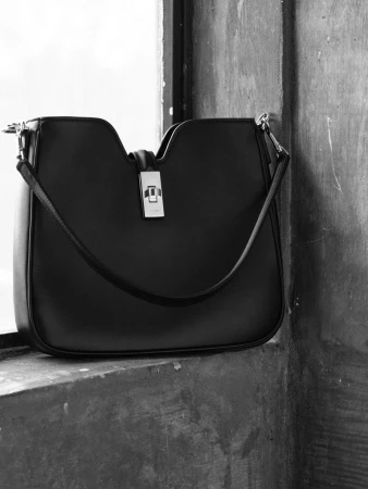 Elegant black leather shoulder bag with a silver clasp, displayed on a textured concrete surface near a window.