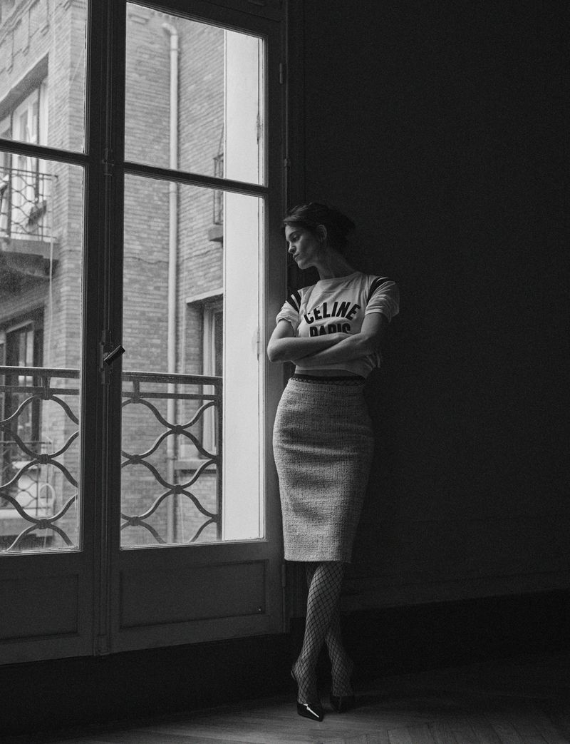 Suzanne Lindon posing indoors next to a tall window in a white t-shirt and tweed skirt, captured in monochrome.