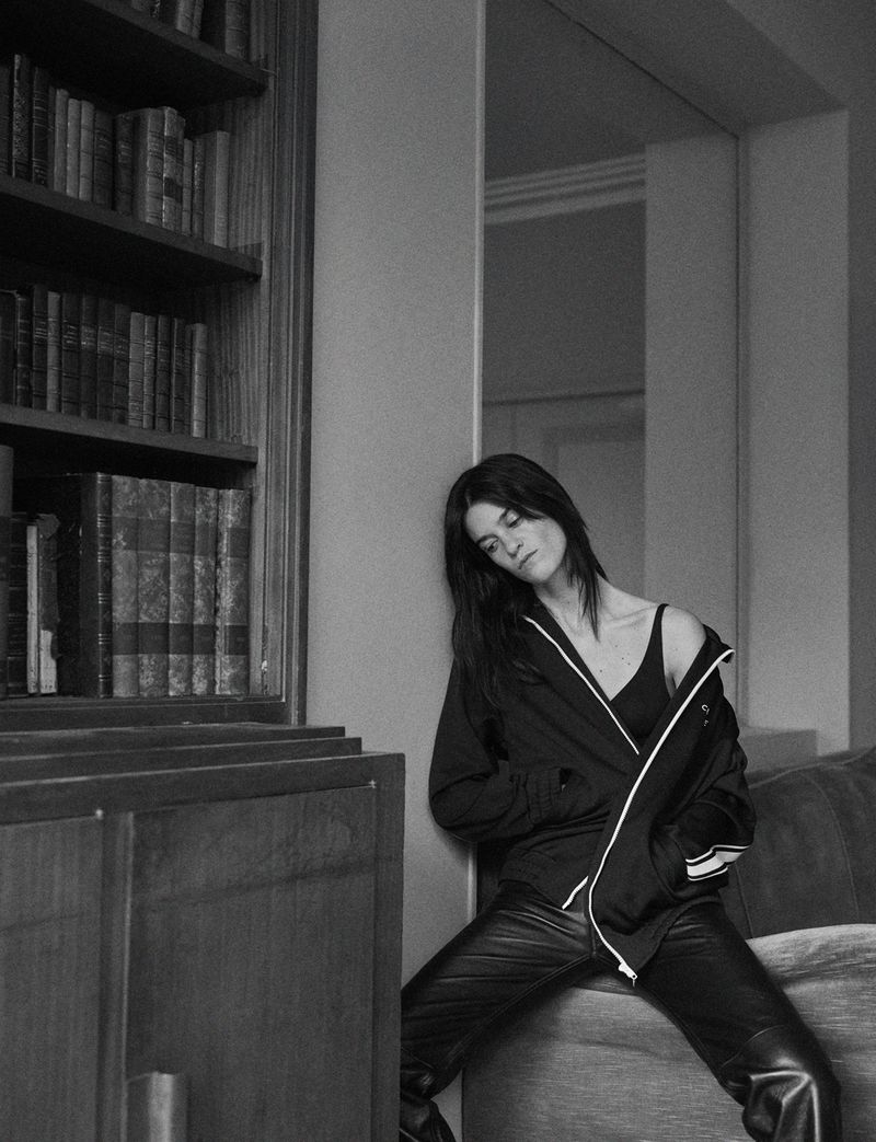 Indoor portrait of a dark-haired woman in a jacket and leather pants leaning against a wall near a bookshelf in black and white.