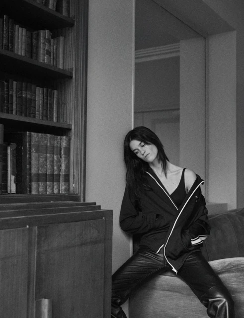 Indoor portrait of a dark-haired woman in a jacket and leather pants leaning against a wall near a bookshelf in black and white.
