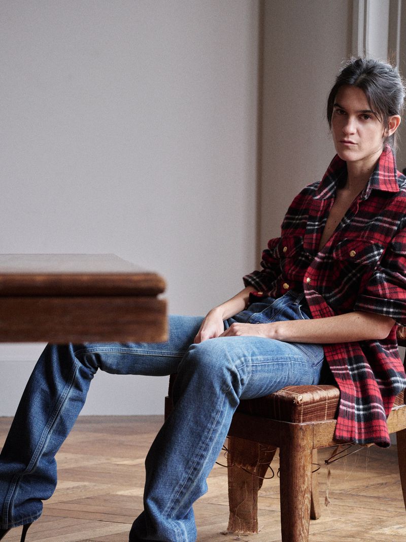 A woman is seated on a wooden stool indoors, wearing a red and black flannel shirt, blue jeans, and black heels in a casual setting.