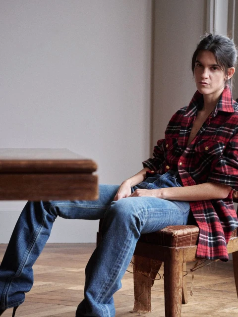 A woman is seated on a wooden stool indoors, wearing a red and black flannel shirt, blue jeans, and black heels in a casual setting.