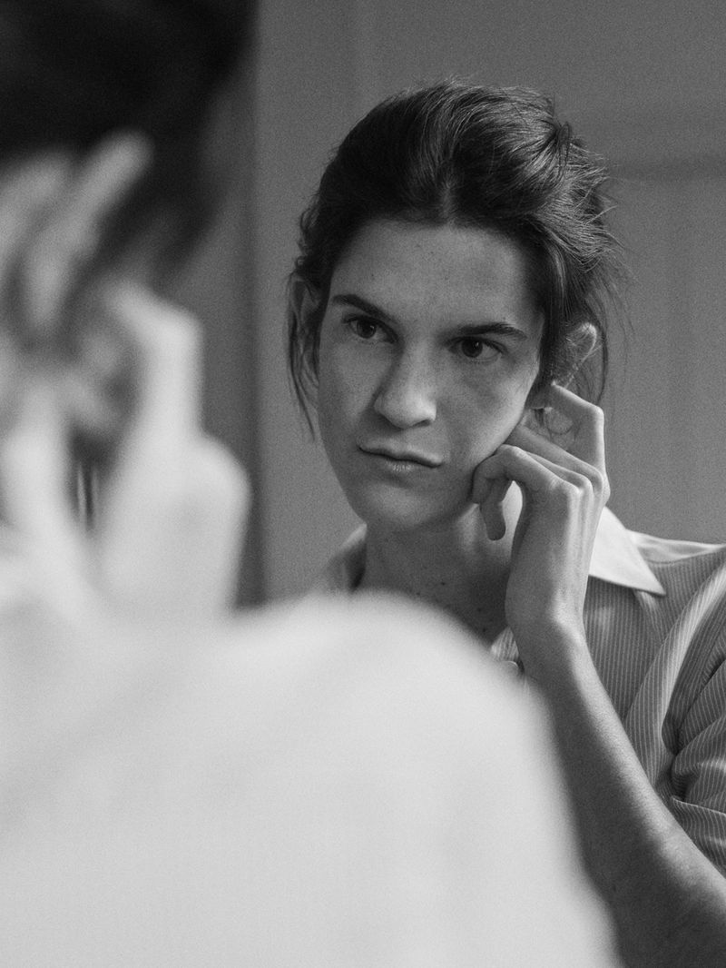 Black and white portrait of a woman looking in the mirror, with hand near face, wearing a button-down shirt indoors with natural light.