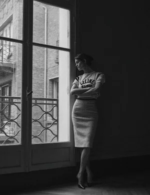 Suzanne Lindon posing indoors next to a tall window in a white t-shirt and tweed skirt, captured in monochrome.