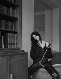Suzanne Lindon leaning against a wall beside a bookshelf in a black jacket and trousers for Heroine Magazine.