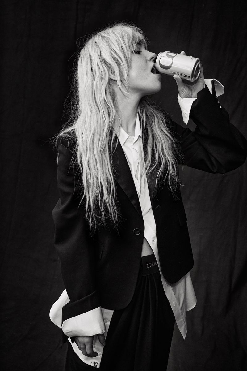 Monochrome portrait of a stylish adult woman drinking from a can, wearing a blazer and oversized shirt against a draped fabric backdrop.