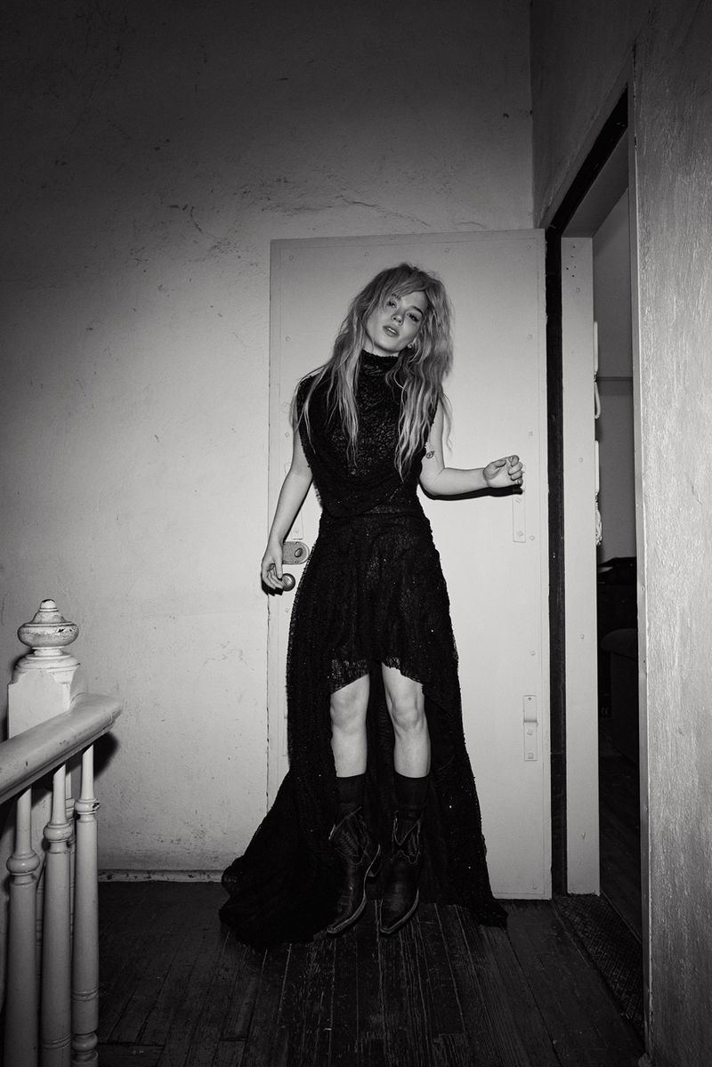 Young woman with blonde hair stands in dark dress and boots, posing in a hallway with an open doorway in a black and white photo.